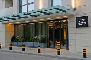 Une entrée d’hôtel moderne avec une façade en verre, une enseigne indiquant « HÔTEL VIKO », et un espace extérieur couvert agrémenté de plantes.