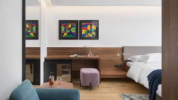 A modern hotel room with a wooden desk, a comfortable sofa, a bed with white linens, and abstract art on the wall.