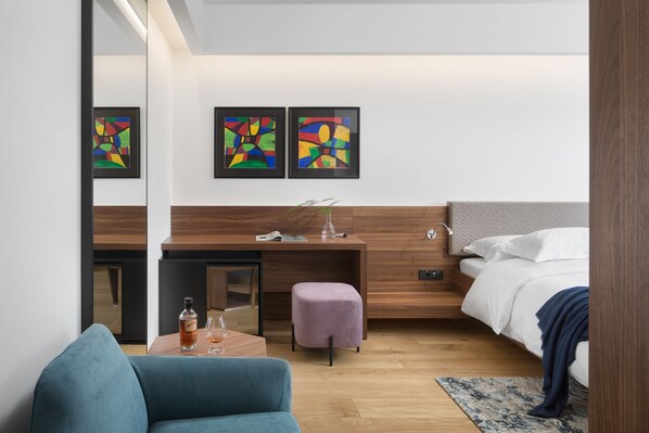 Una habitación de hotel moderna con un escritorio de madera, un sofá cómodo, una cama con ropa blanca y arte abstracto en la pared.