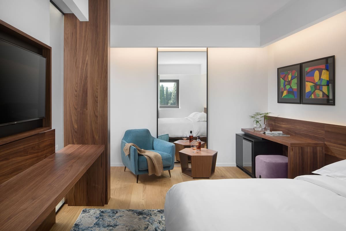 A modern hotel room with a large bed, a seating area with a blue armchair, a small desk, and a television.