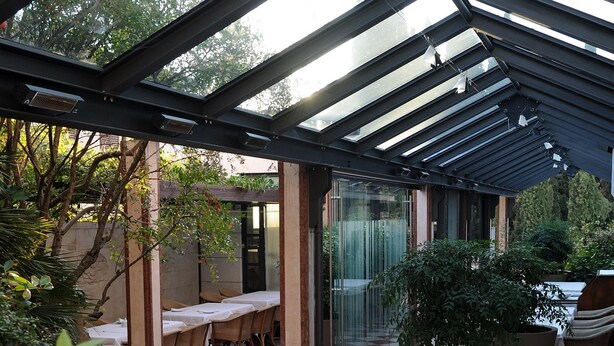 An outdoor patio with tables and chairs, covered by a glass roof, surrounded by greenery. - Hotel & Restaurant Milan (Pula)