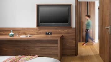 Una habitación de hotel moderna con un televisor de pantalla plana grande montado en la pared, un escritorio de madera con un florero y un vaso, y una cama con una manta estampada floral.