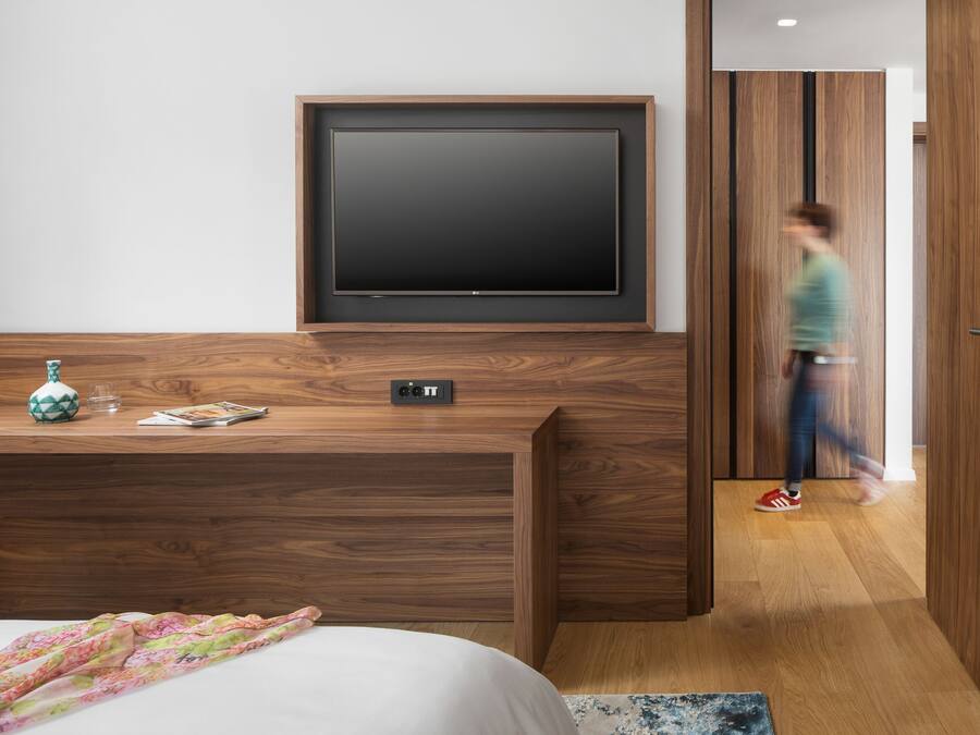 Una habitación de hotel moderna con un televisor de pantalla plana grande montado en la pared, un escritorio de madera con un florero y un vaso, y una cama con una manta estampada floral.