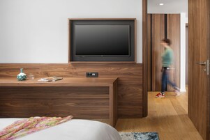 Una habitación de hotel moderna con un televisor de pantalla plana grande montado en la pared, un escritorio de madera con un florero y un vaso, y una cama con una manta estampada floral.