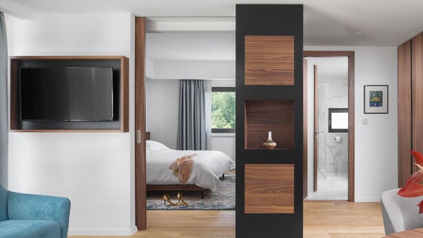 Una habitación de hotel moderna con un televisor de pantalla plana grande, una cama con manta gris, un sofá azul y una pared de acento de madera.