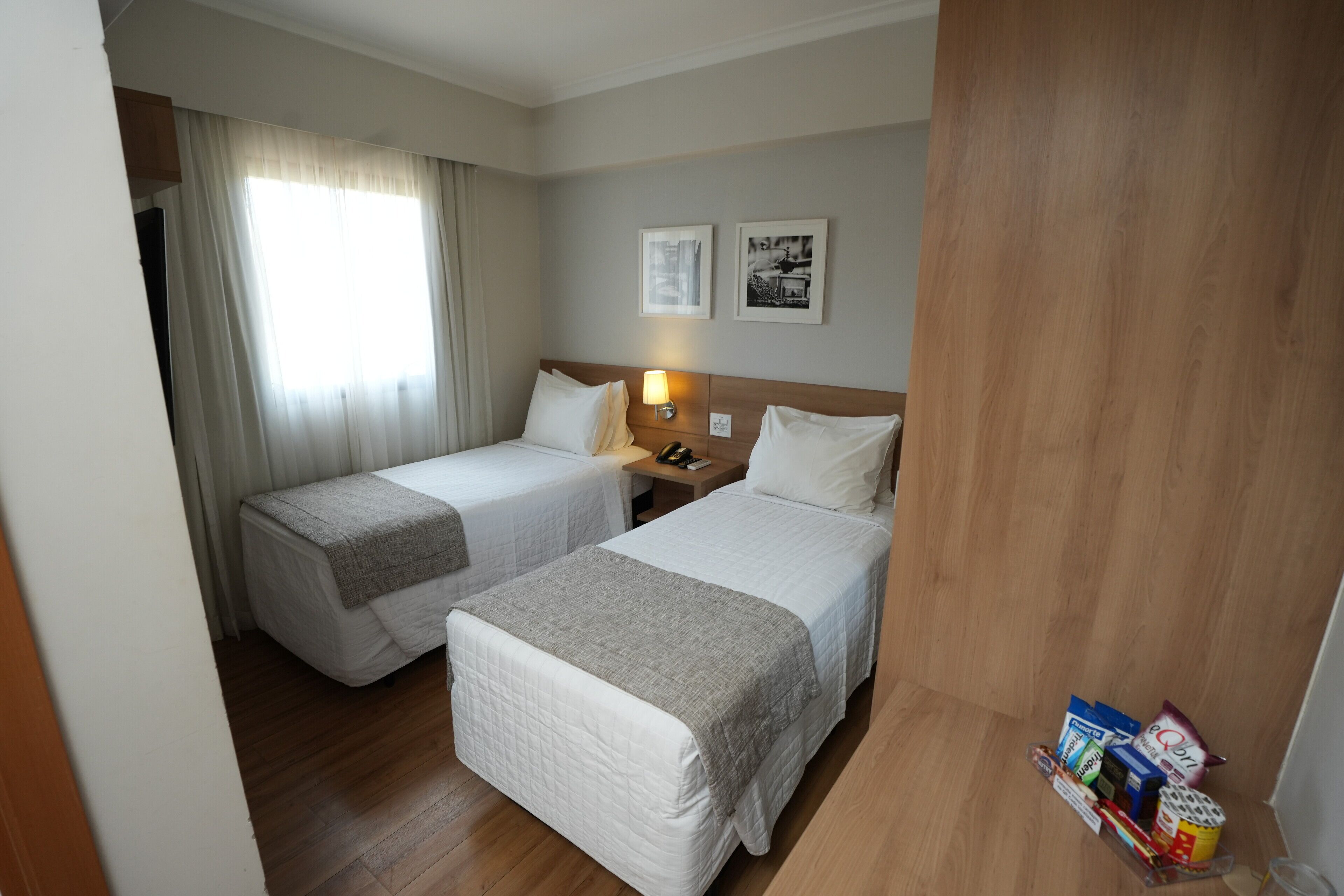 A hotel room with two single beds, a wooden floor, a nightstand with a lamp, and a small table with snacks.