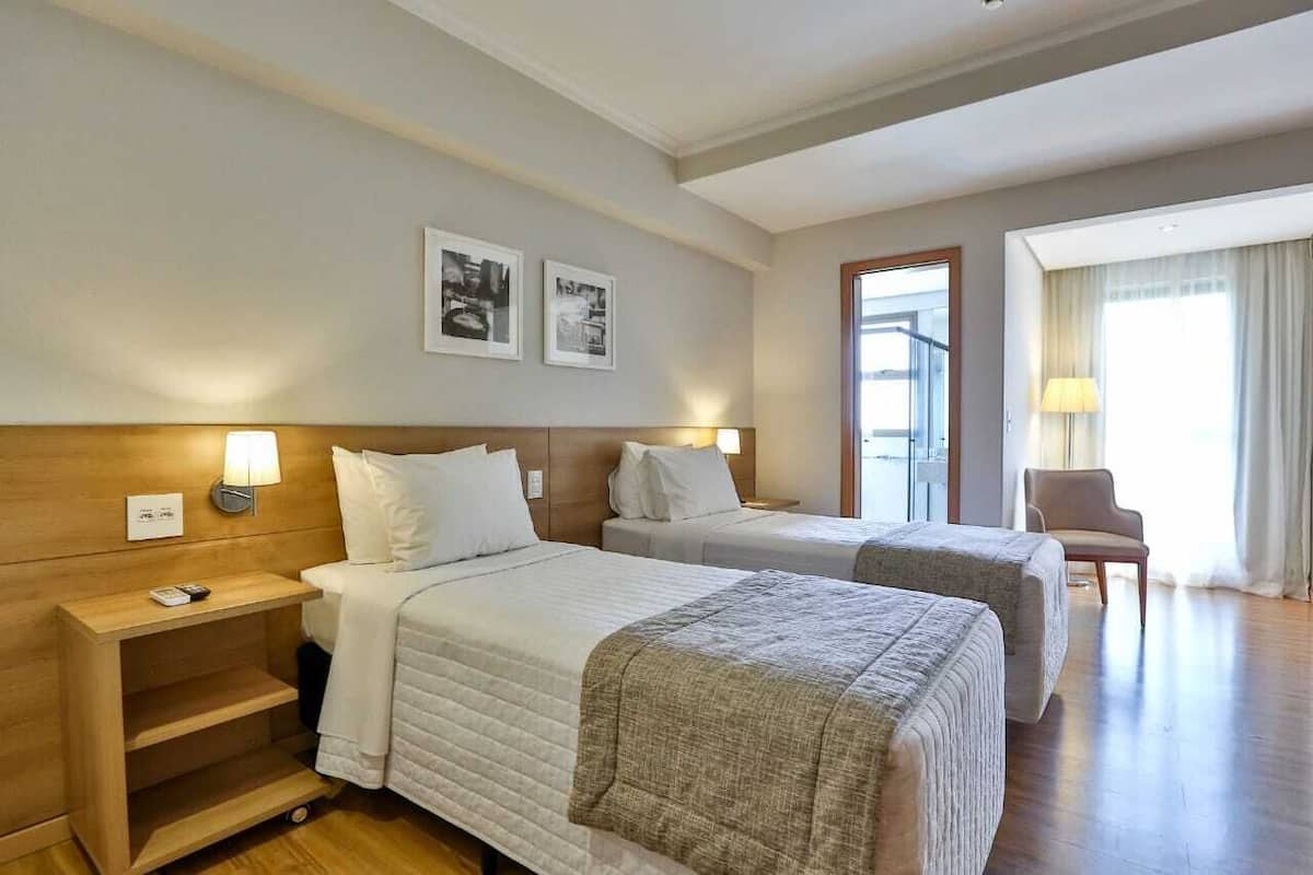 Habitación de hotel con dos camas, cabeceros de madera, lámparas de noche, una mesita con un control remoto y una silla cerca de una ventana con cortinas.