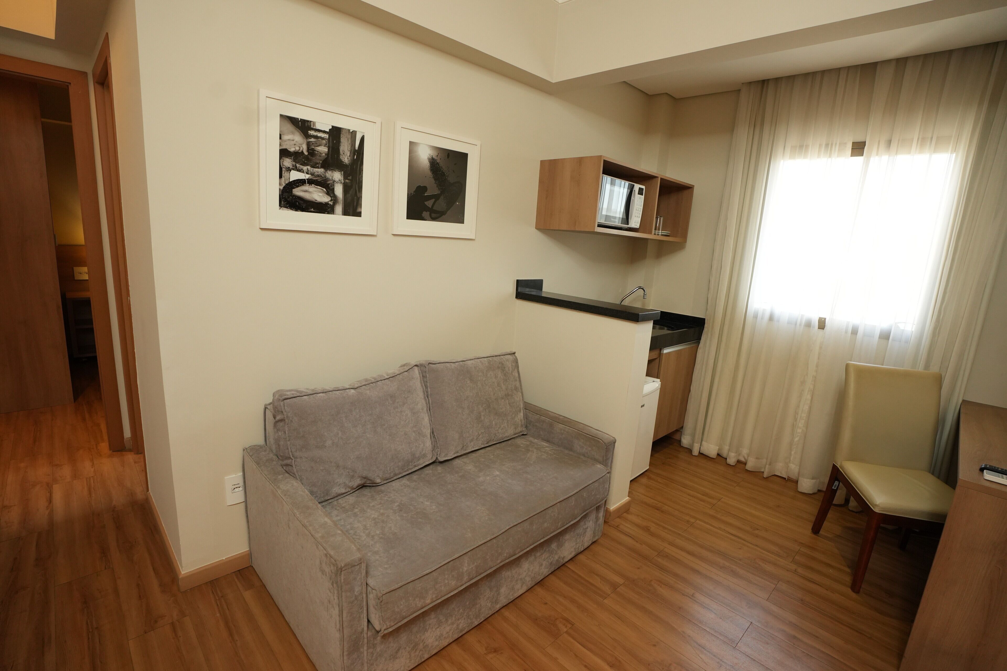 A living room with a sofa, a chair, a small kitchen area with a microwave, and a window with curtains.