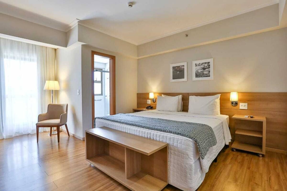 Una habitación de hotel con una cama grande, mesitas de noche, una silla y un banco.