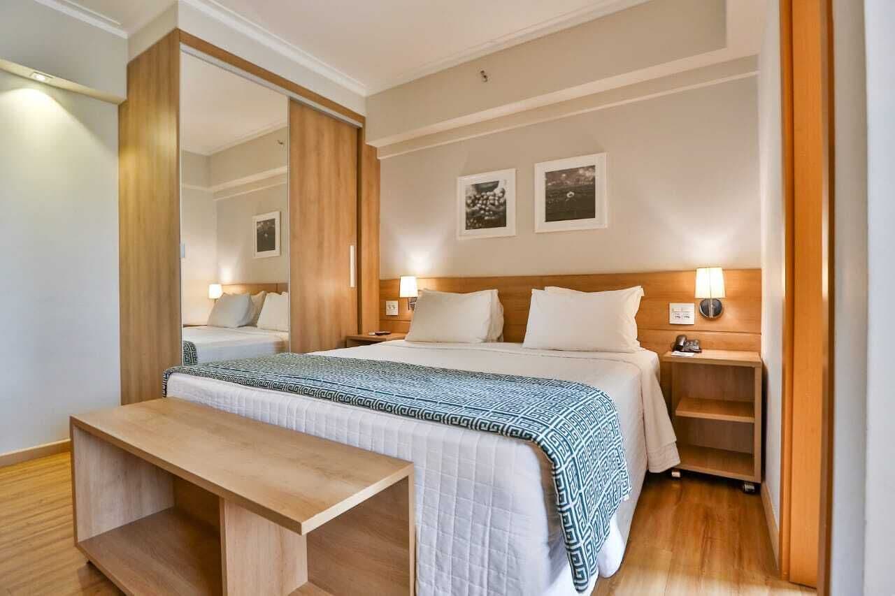 Una habitación de hotel con una cama, mesitas de noche, un banco y un espejo.