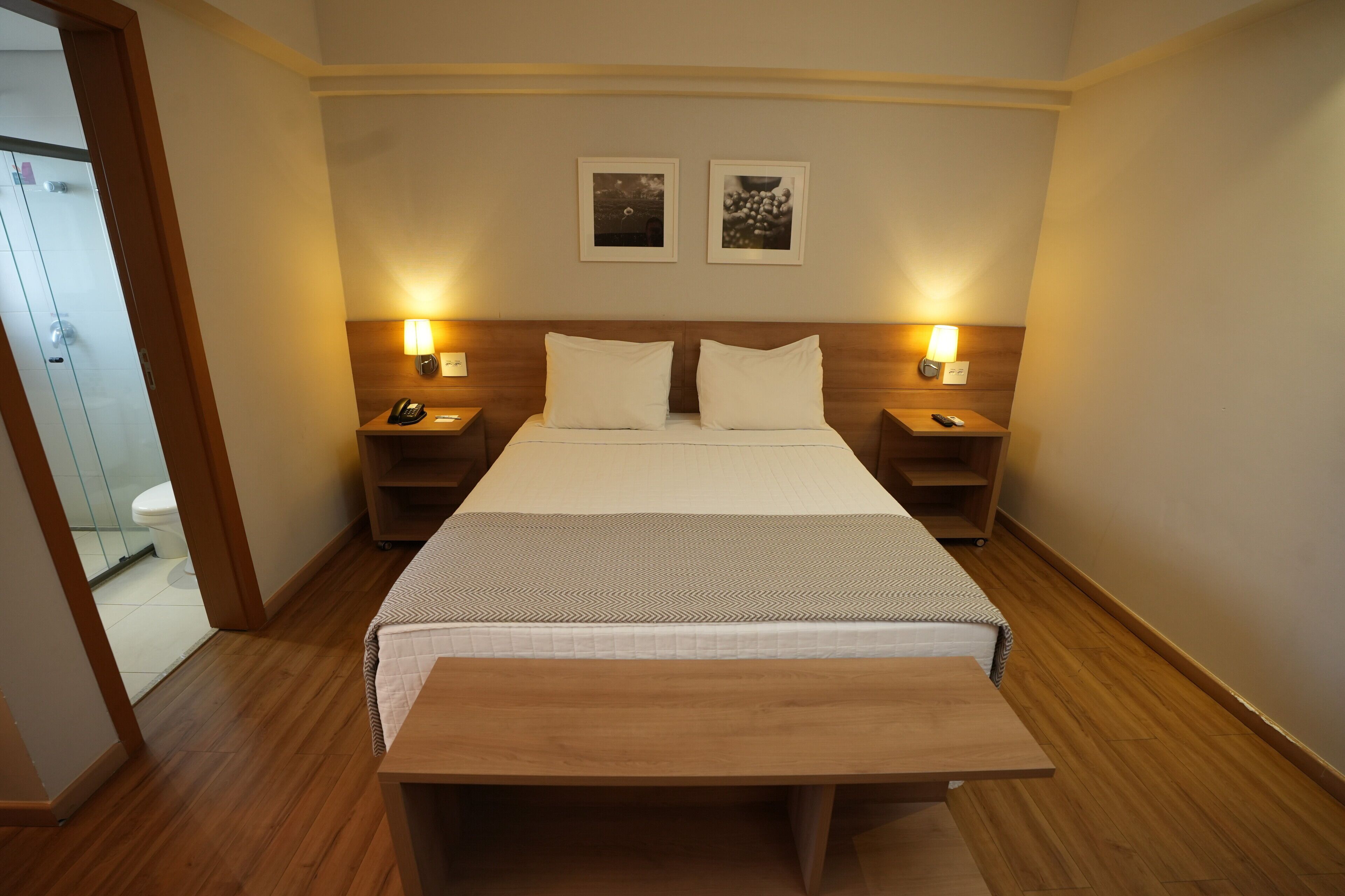 Ein Hotelzimmer mit einem Bett, Nachttischen, einem kleinen Tisch, einer hölzernen Bank, zwei Wandlampen und zwei gerahmten Bildern.