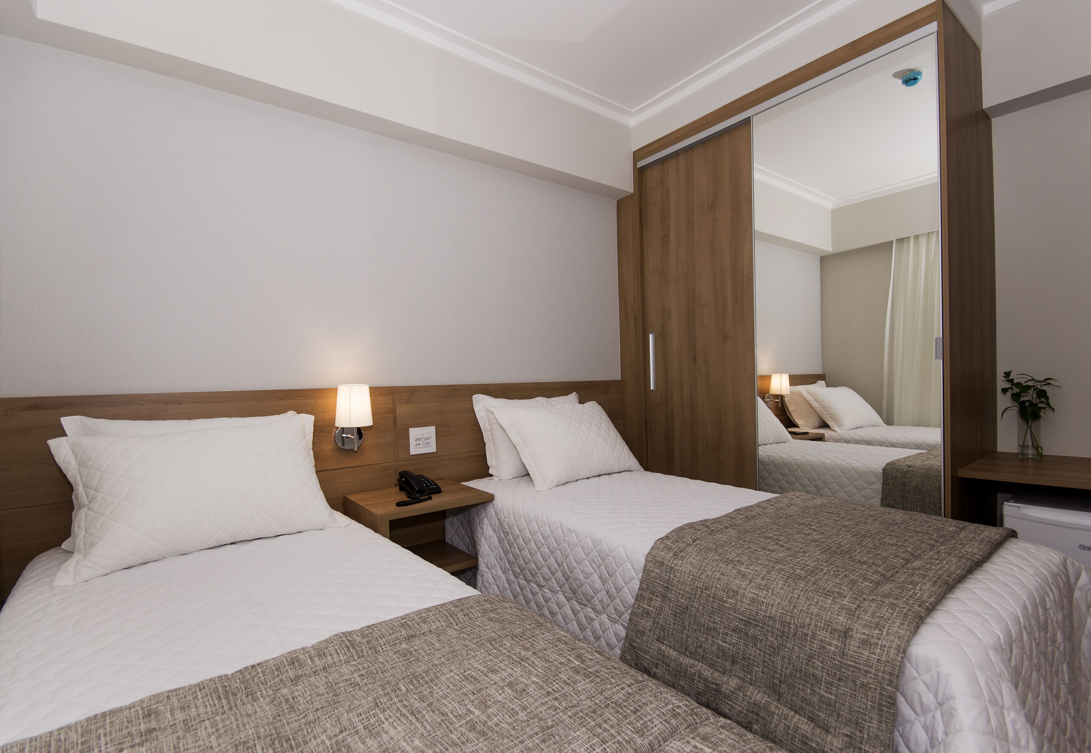 Habitación de hotel con dos camas, cabecera de madera, lámparas de noche, mesitas de noche, un espejo y un pequeño escritorio con un florero.