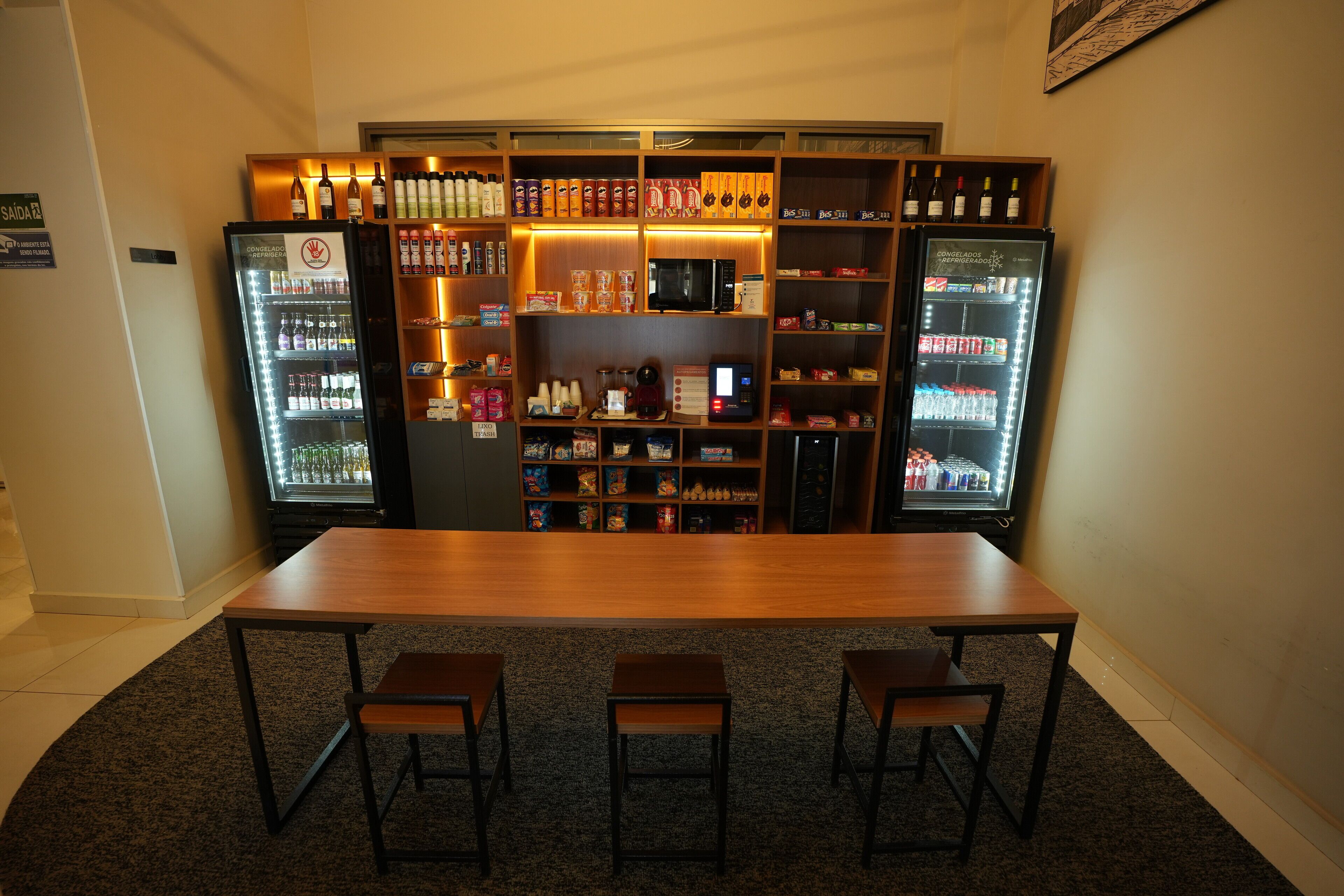 Una zona de bar con una mesa de madera larga y tres sillas, rodeada de estanterías llenas de bebidas y aperitivos.