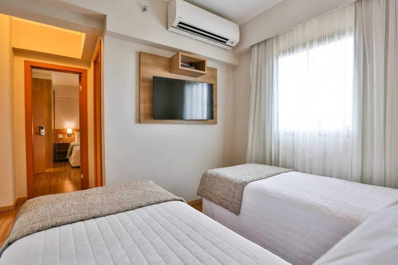 A hotel room with two beds, a mounted TV, and an air conditioning unit.