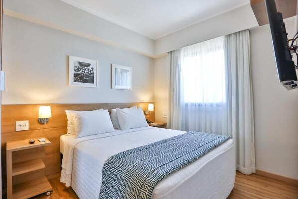 Quarto de hotel com cama, mesas de cabeceira, luminária e janela com cortinas.