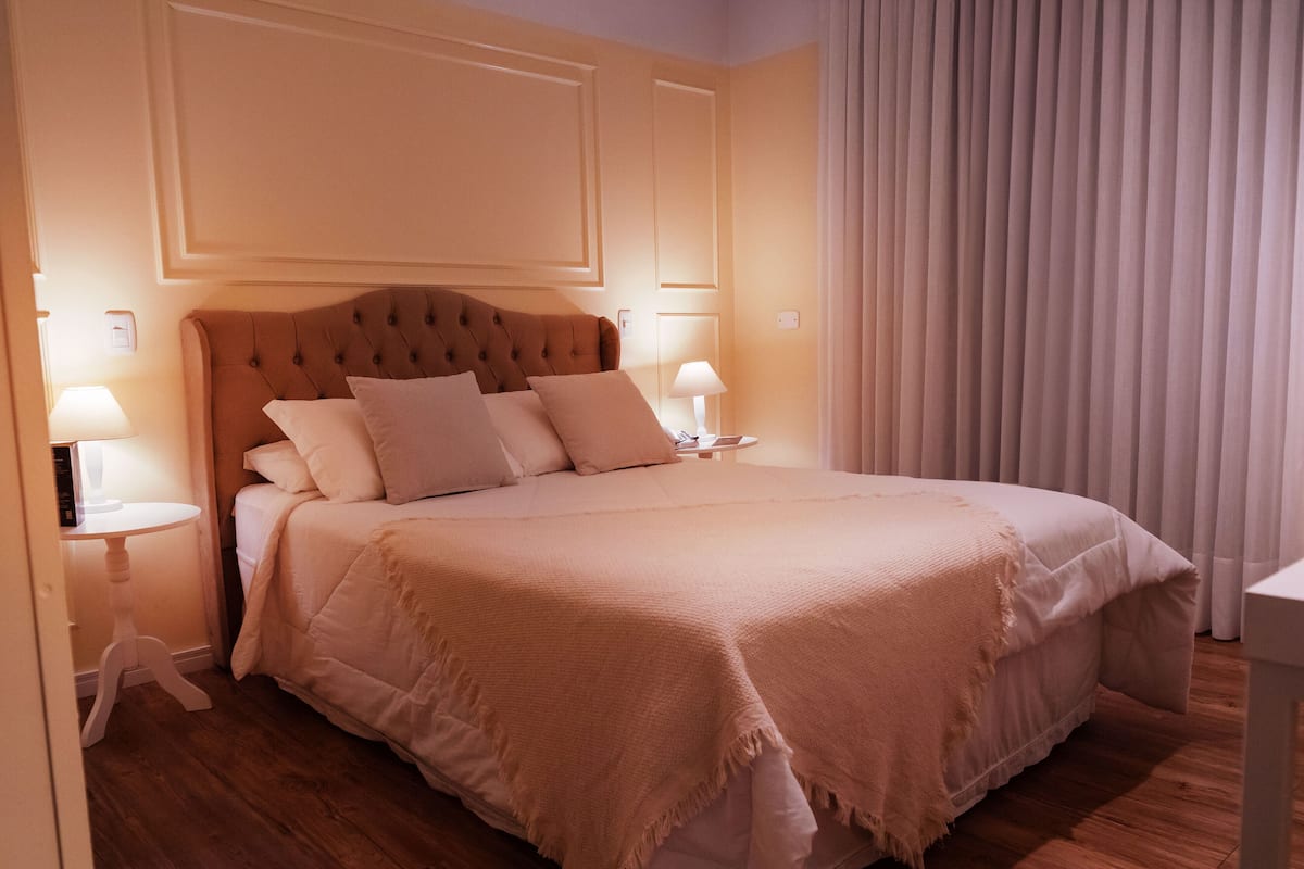 Una cama bien tendida con un cabecero capitonado, dos lámparas de noche y un ventanal con persianas verticales.