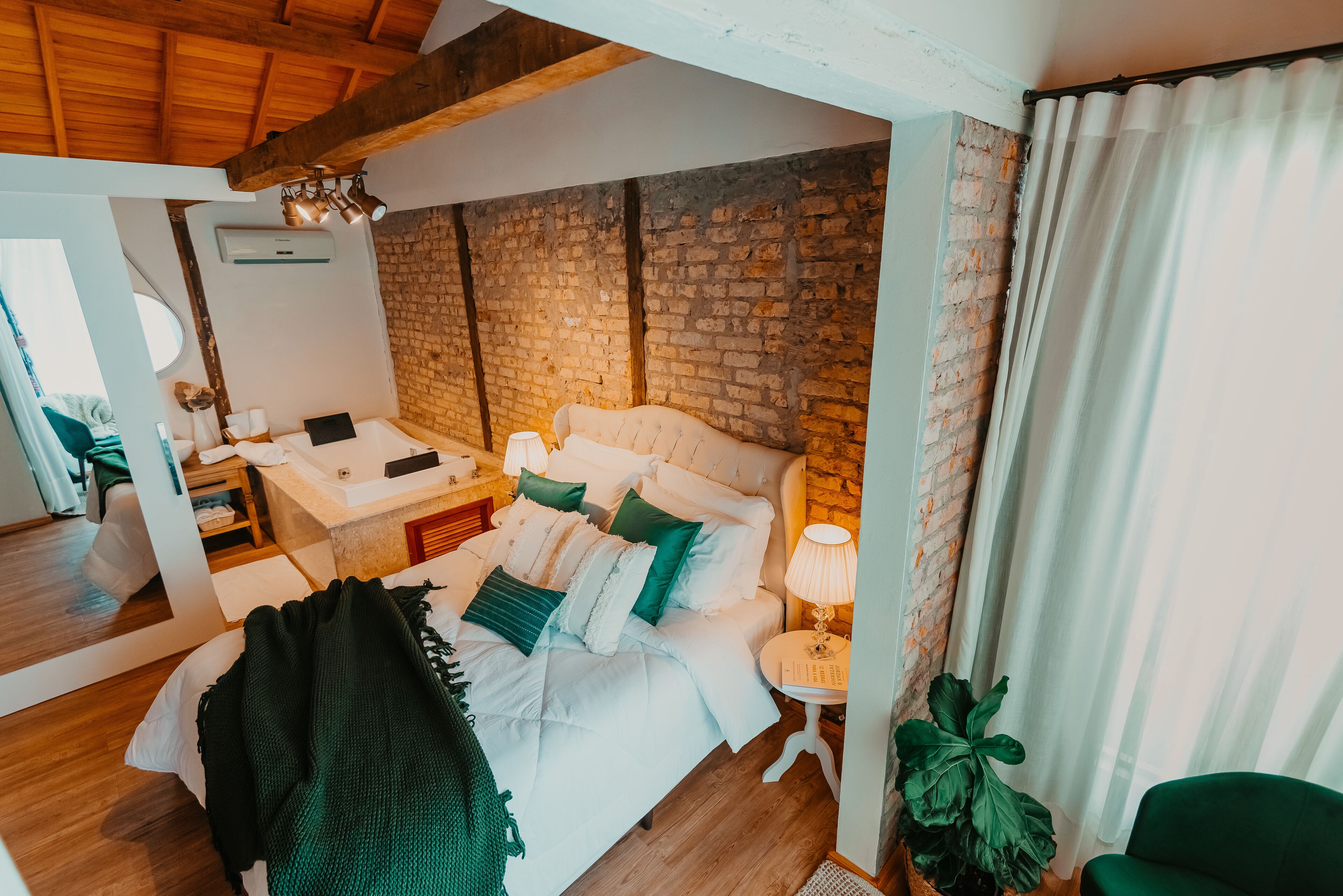A bedroom with a brick wall, a bed with white bedding, a bedside table with a lamp, and a green chair.