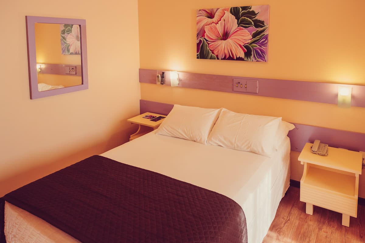 Una habitación de hotel con una cama, una mesita de noche con un teléfono, una lámpara y un cuadro en la pared.