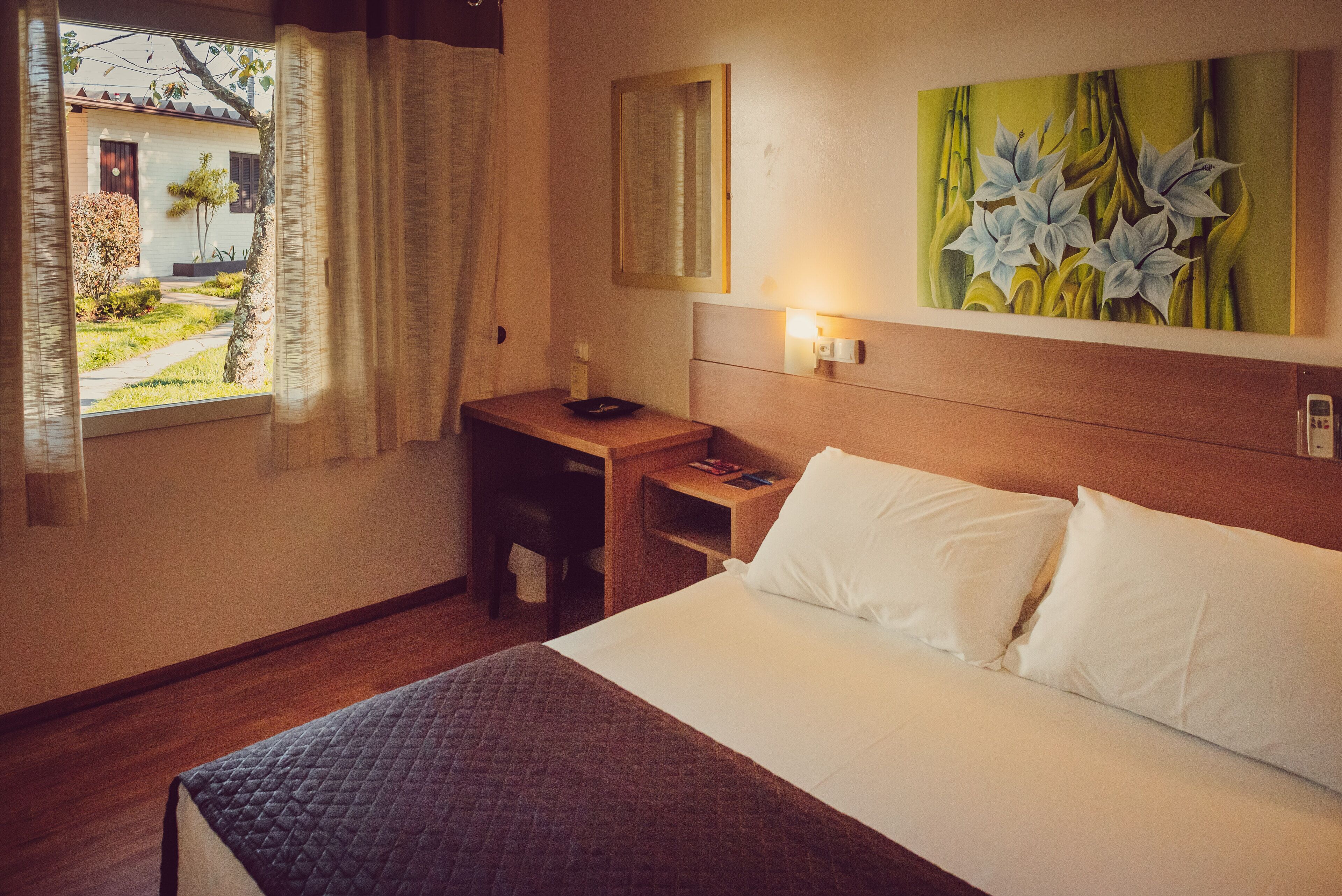 Une chambre d’hôtel avec un lit, une table de chevet, une commode, une fenêtre avec des rideaux, un tableau au mur et une vue sur l’extérieur.