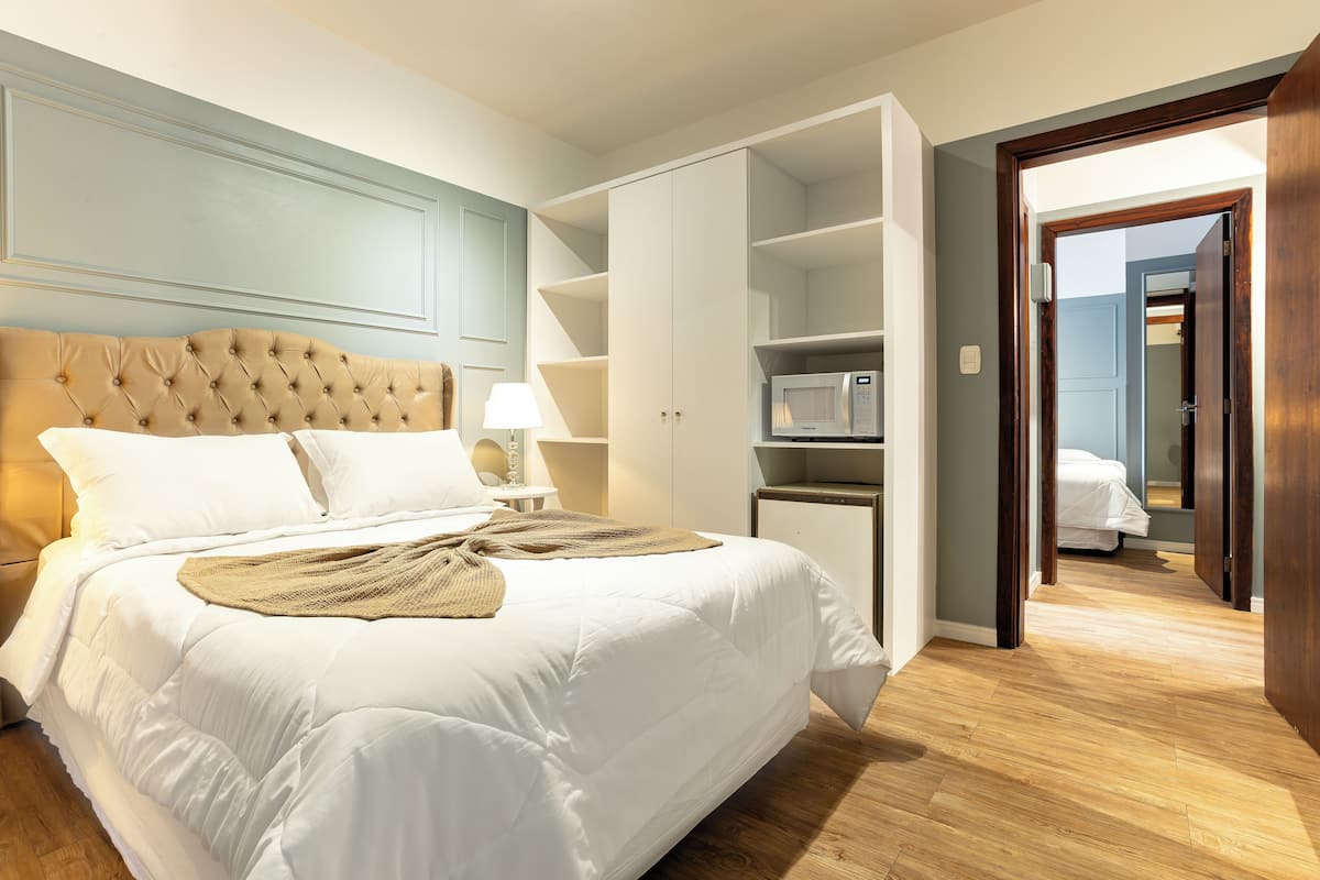 Una cama bien hecha con cabecera capitonada, una lámpara de noche y un microondas sobre un mueble de madera.