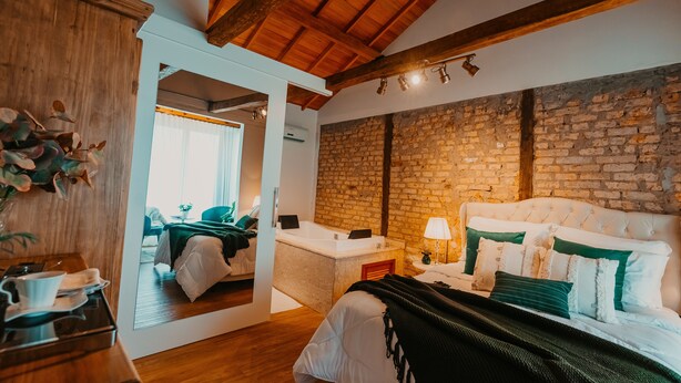 Un dormitorio con pared de piedra, vigas de madera en el techo, una cama blanca y un baño con lavamanos y espejo blancos.