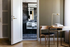 A modern bathroom with a mirror, sink, and towel rack. A wooden chair and desk with a lamp are positioned near a window.