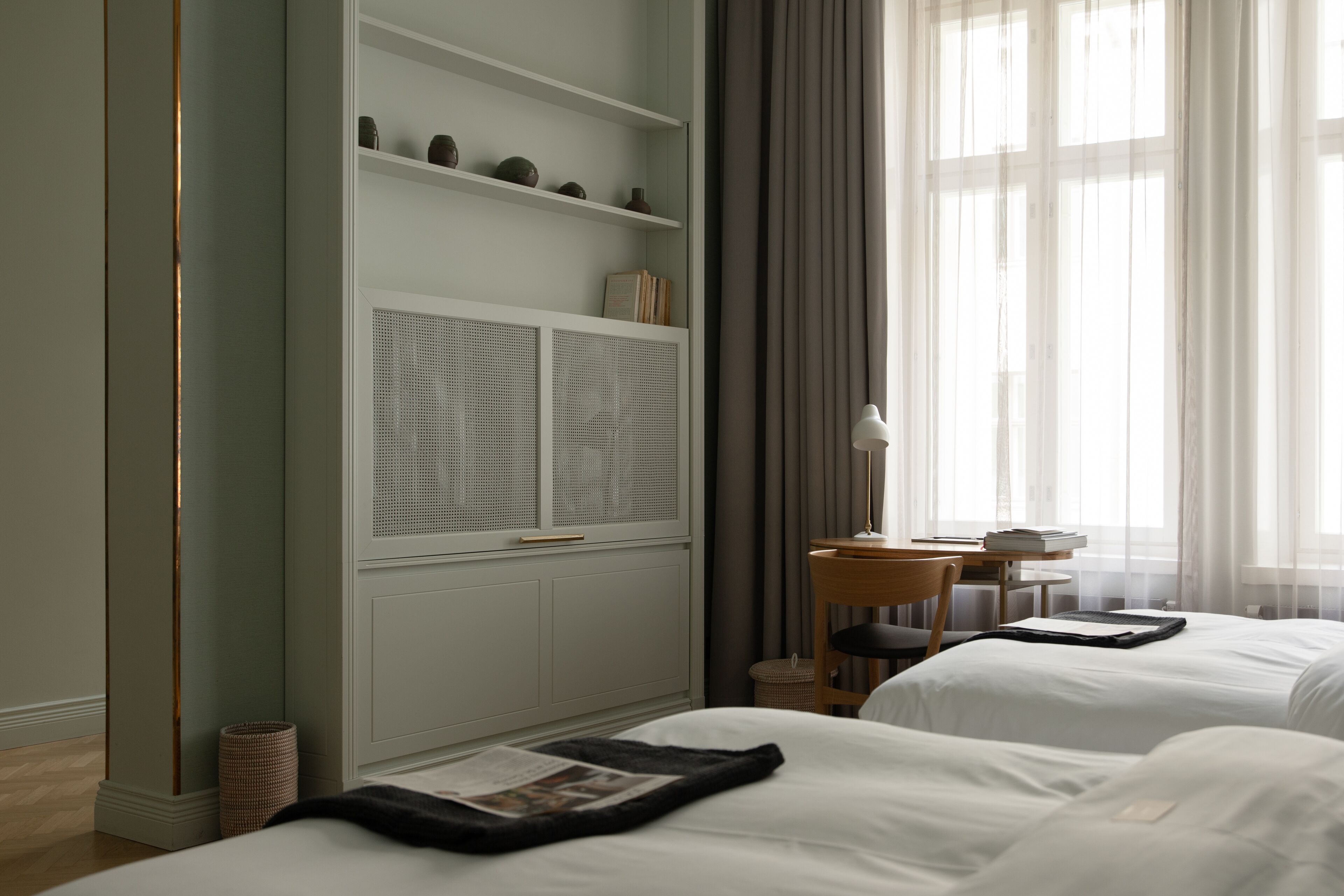A hotel room with two beds, a bookshelf, a desk with a lamp, and a window with curtains.
