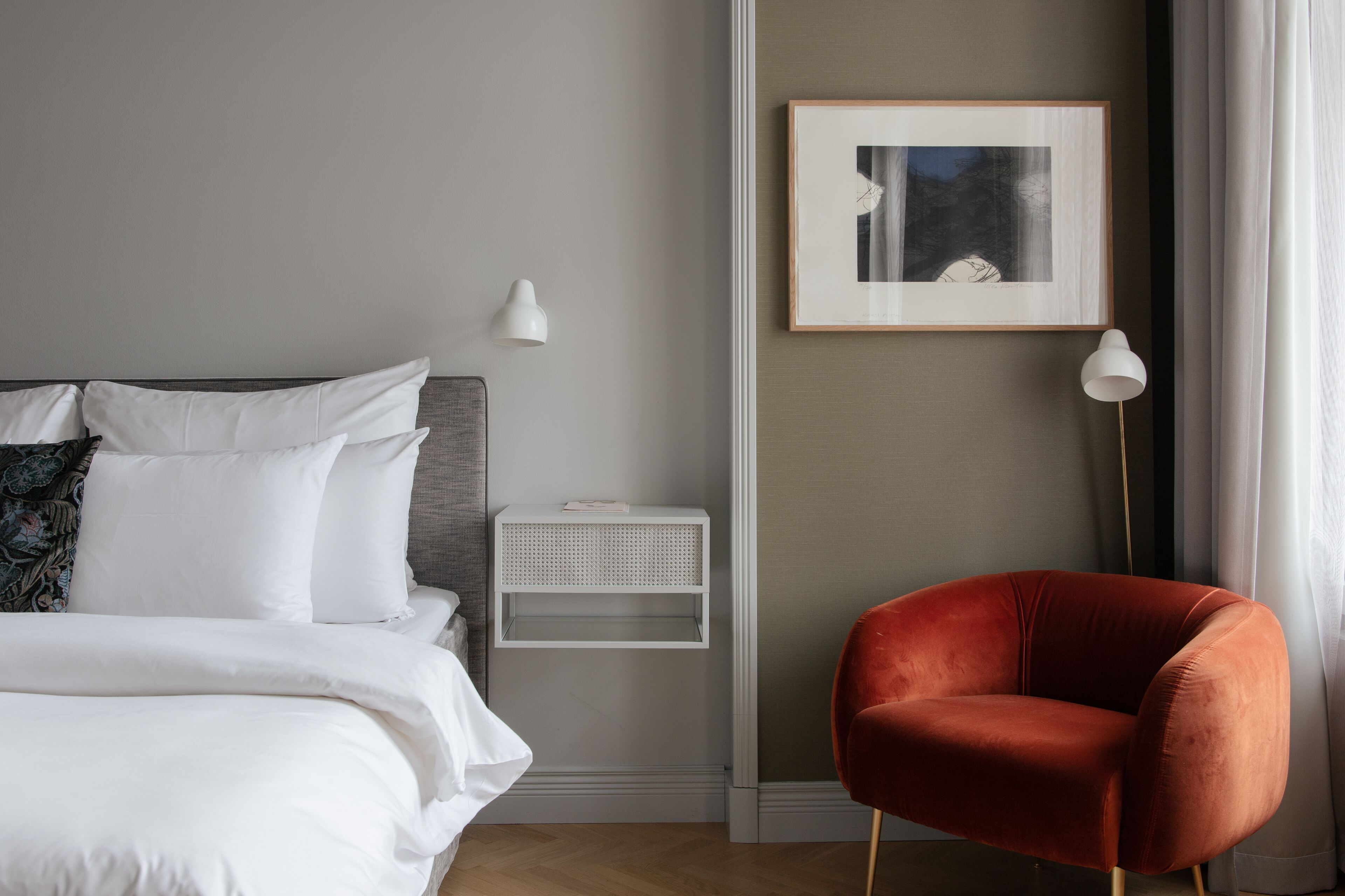 Un dormitorio ordenado con cama, una silla roja, una mesita de noche, una lámpara de pared y un cuadro enmarcado.