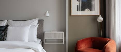Un dormitorio ordenado con cama, una silla roja, una mesita de noche, una lámpara de pared y un cuadro enmarcado.