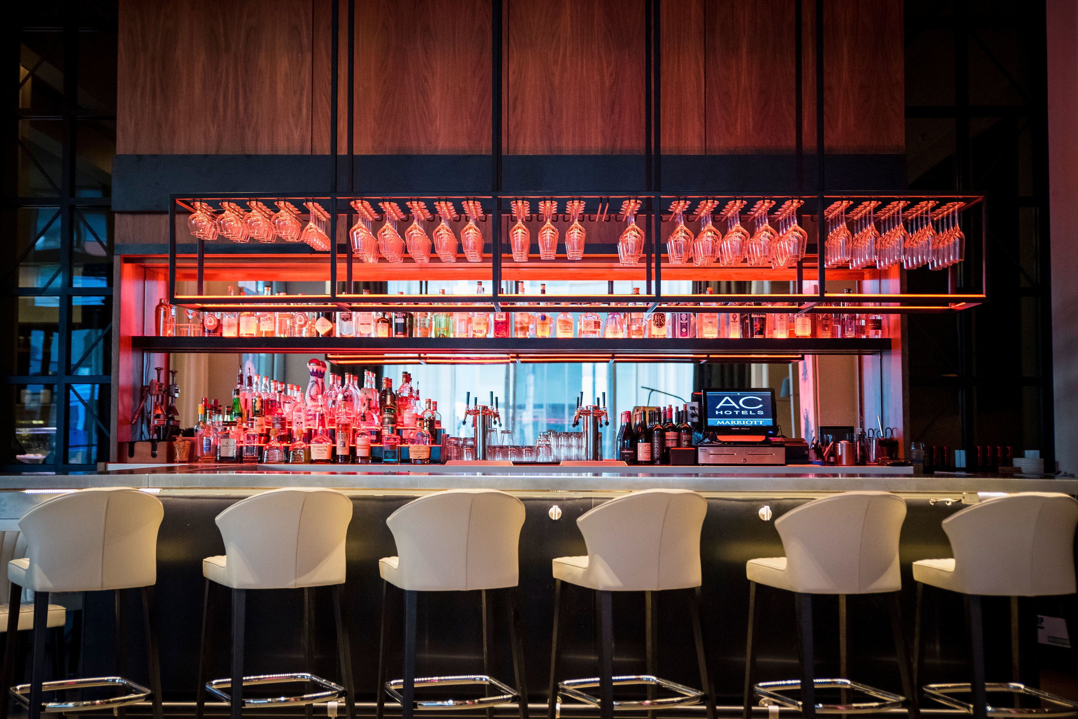 A bar area with multiple shelves of bottles and glasses, a digital screen displaying the hotel name, and several white bar stools.