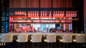 Een bar met meerdere flessen en glazen achter de bar, een scherm dat de naam van het hotel toont, en verschillende witte barkrukken.