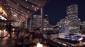 An outdoor seating area with a fire feature, overlooking a cityscape with illuminated buildings.