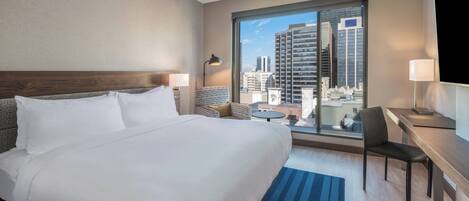 Habitación de hotel con una cama grande, un escritorio, una silla y vistas a la ciudad a través de un ventanal amplio.
