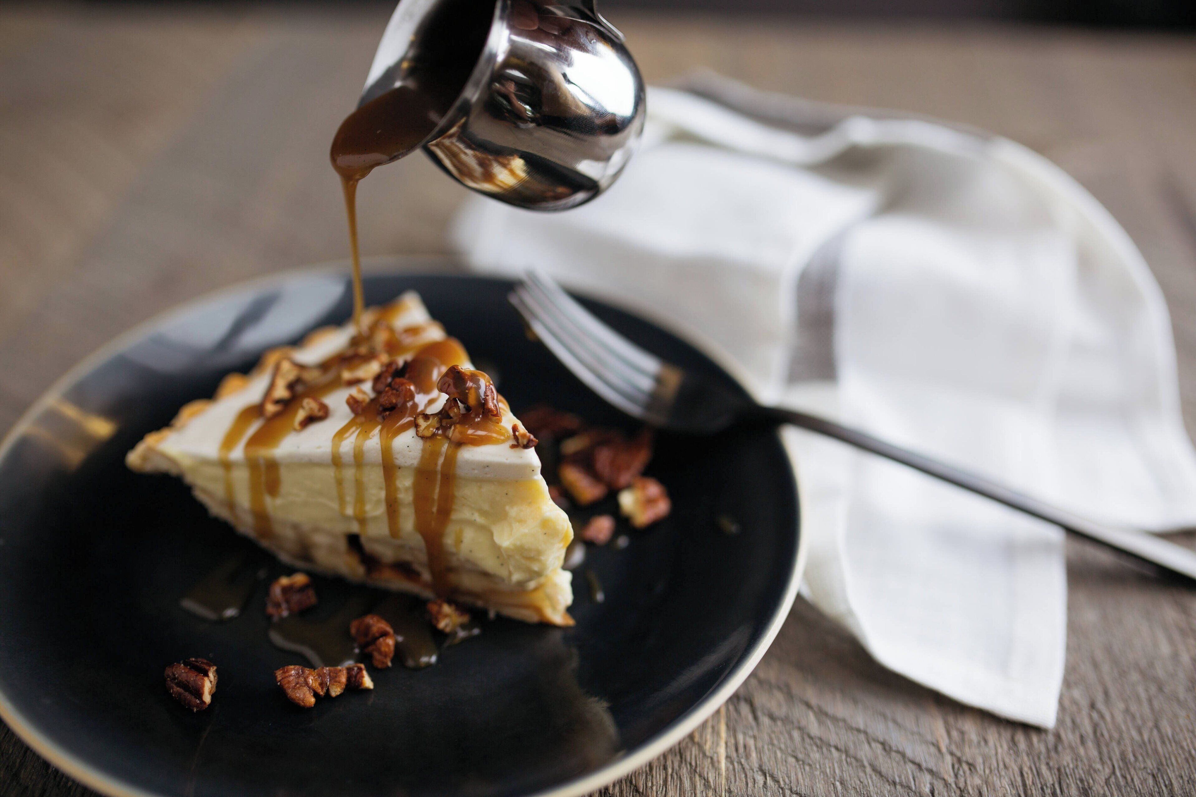 A slice of cheesecake with caramel sauce and pecans on a black plate.