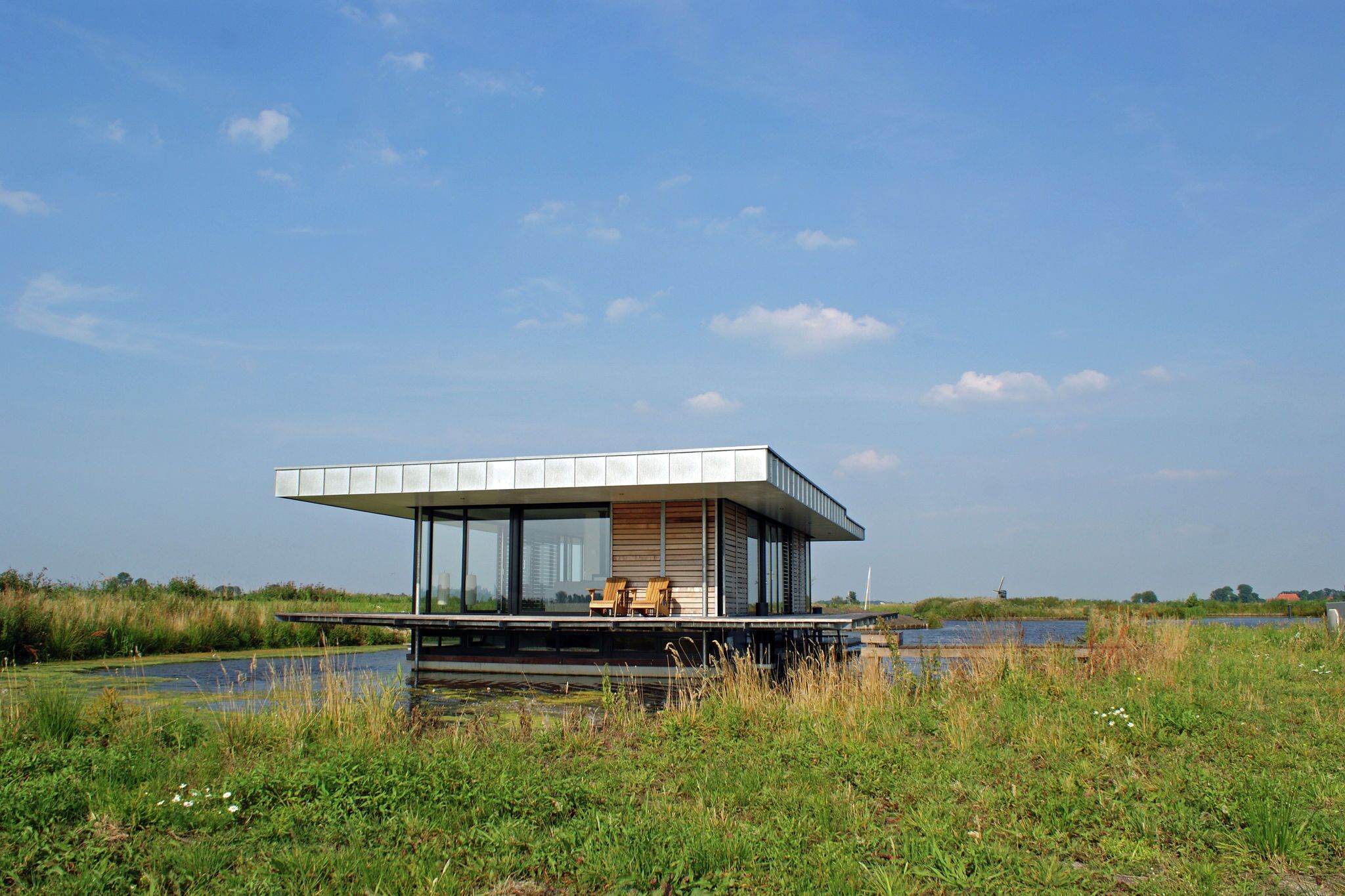 Une péniche moderne dotée d’un toit plat, de grandes fenêtres et d’une terrasse avec des chaises.
