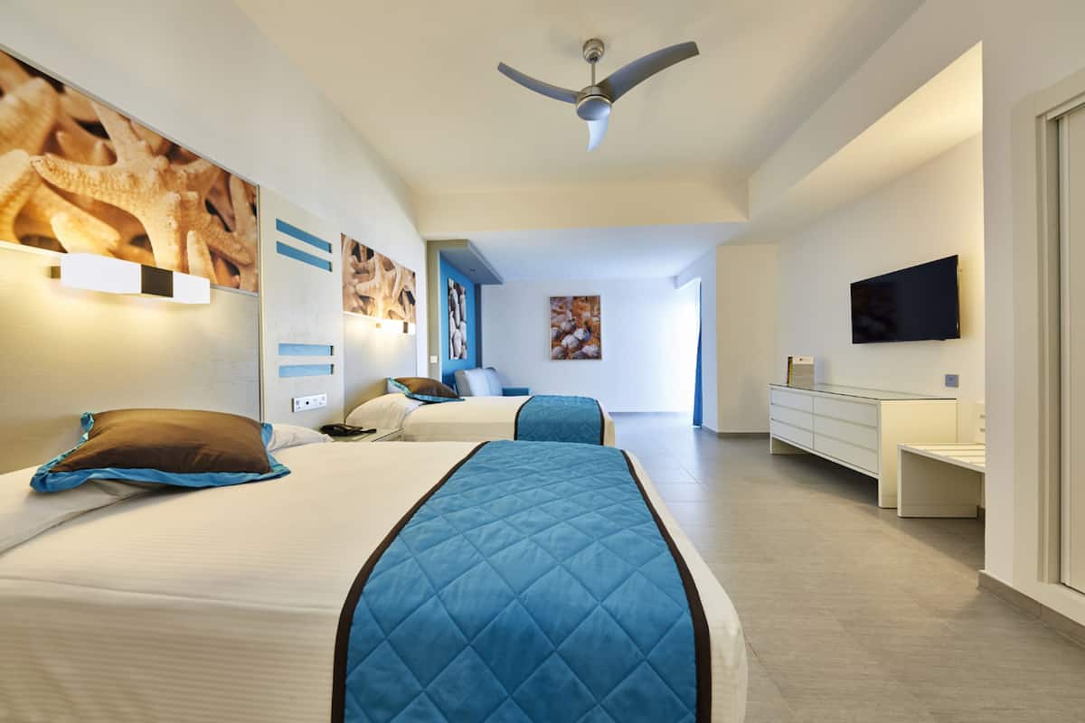 A modern hotel room with two beds, a ceiling fan, artwork on the walls, a flat-screen TV, and a large window.