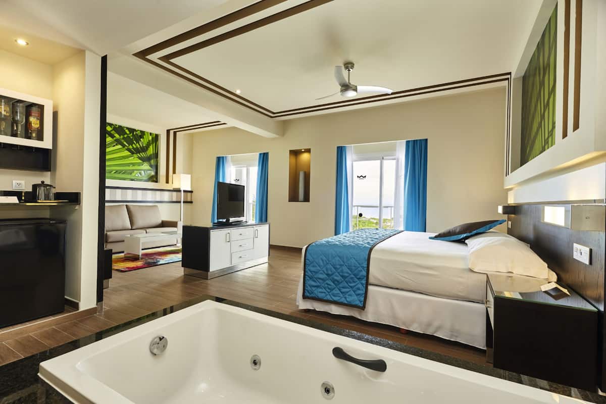 A modern hotel room with a large bed, a separate bathroom with a bathtub, a dining area, and a view of the outdoors.
