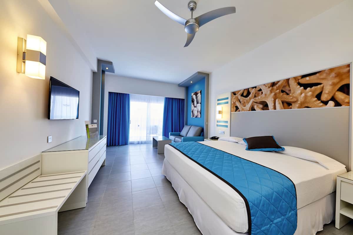 A modern hotel room with a bed, a desk, a chair, a TV, and a ceiling fan.