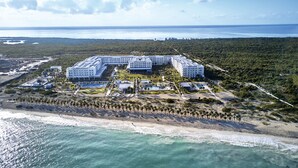 Une vue aérienne d’un complexe hôtelier comprenant plusieurs bâtiments, une plage et une forêt dense en arrière-plan.
