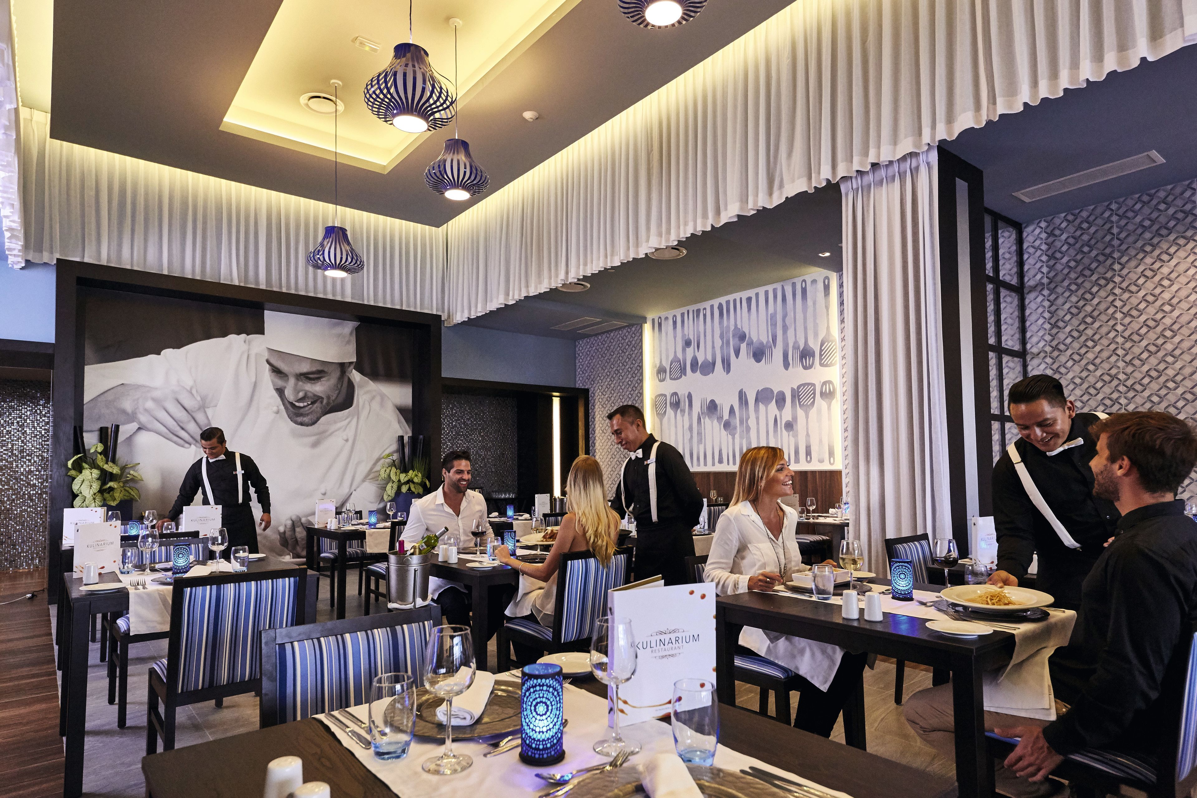 A restaurant with tables set for dining, waitstaff attending to guests, and a large mural on the wall.