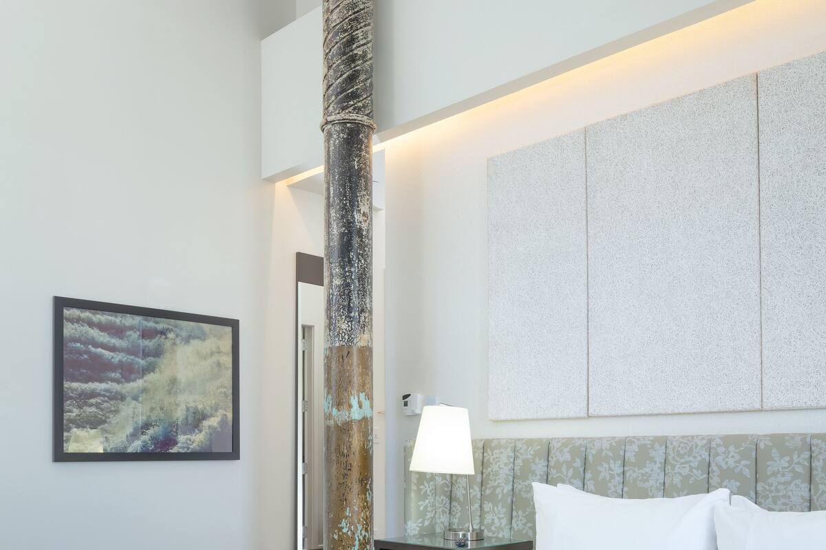 Una habitación de hotel con una cama grande, una mesita de noche, una lámpara, un cuadro enmarco y un pilar decorativo.