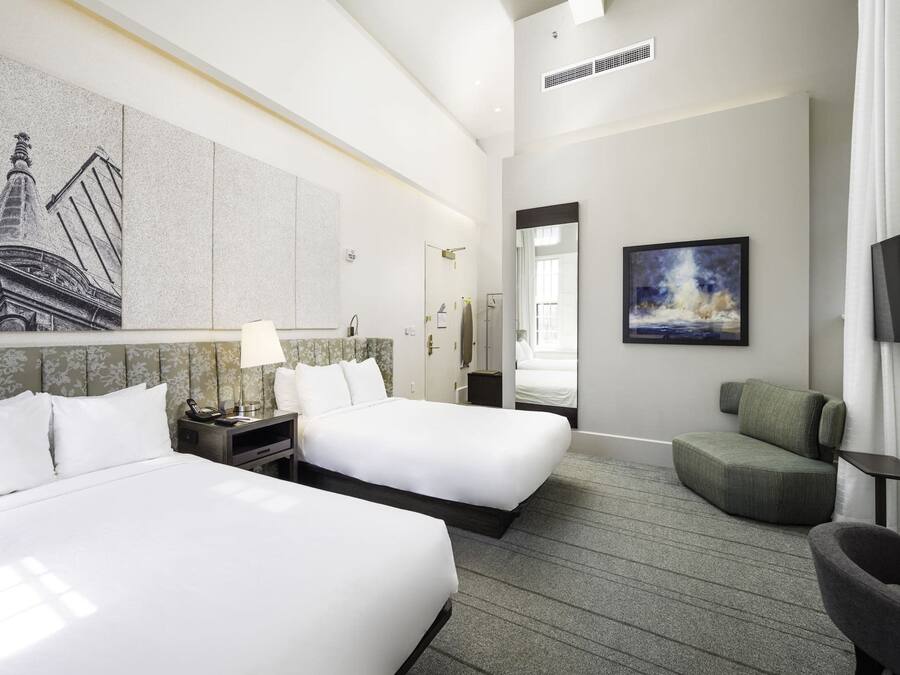 A hotel room with two beds, a green armchair, a large painting, and a view of a building.