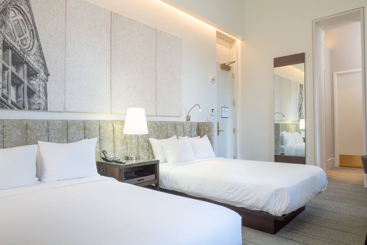Habitación de hotel con dos camas, una mesita de noche con lámpara, un espejo grande y una obra de arte en la pared.