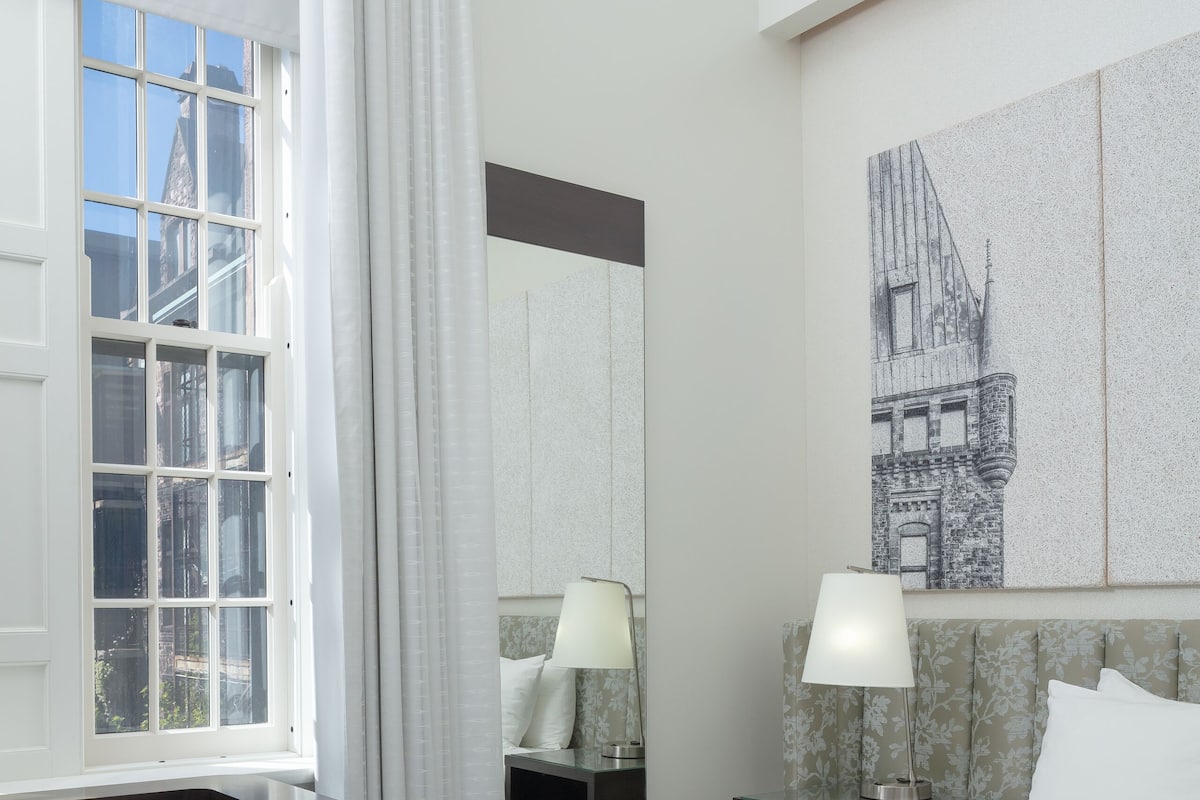 Habitación de hotel con cama, escritorio, silla, mesita de noche, lámpara, ventana con cortinas y un cuadro colgado en la pared.