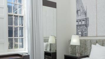 Habitación de hotel con cama, escritorio, silla, mesita de noche, lámpara, ventana con cortinas y un cuadro colgado en la pared.