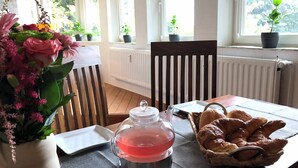 Una mesa con una tetera, un plato y una cesta de cruasanes. Hay plantas en macetas en el alféizar de la ventana y una silla al fondo.