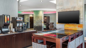 Un área de bar moderna con una televisión de pantalla plana, máquinas de café y un espacio de estar con sillas estampadas.