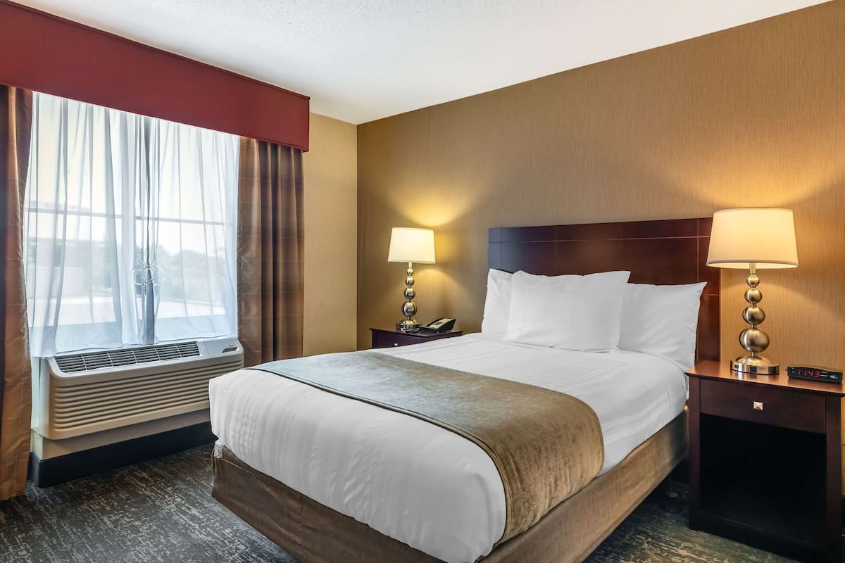 A hotel room with a large bed, two bedside tables with lamps, a window with curtains, and an air conditioning unit.