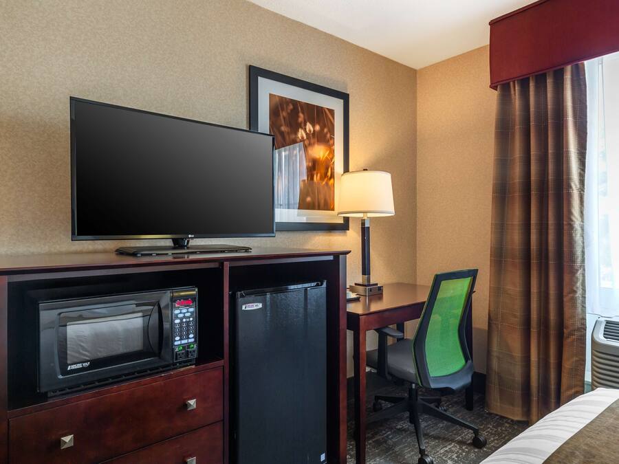 A hotel room with a TV, microwave, mini-fridge, desk, chair, lamp, and a window with curtains.