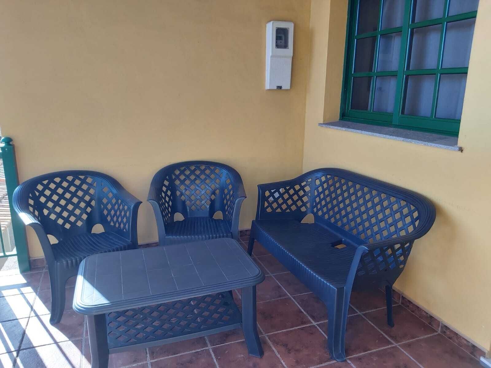 A small outdoor seating area with a table and three chairs, a window with green frames, and a wall-mounted air conditioning unit.